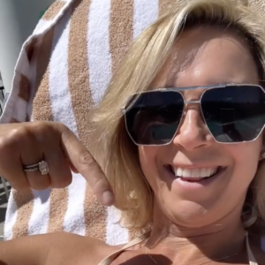 A woman with blonde hair and sunglasses is smiling while pointing downward. She is lying on a lounge chair covered with a striped towel. The background is bright and sunny.